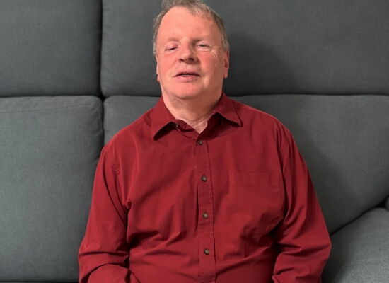 Bruno Etzenberger sitzt in einem grauen Sessel. Er trägt ein rotes Hemd und hat die Hände gefaltet.