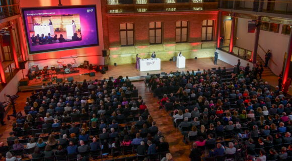 Von schräg hinten wird über das festlich gekleidete Publikum hinweg die Bühne fotografiert. Vorne steht Moderatorin Nadine Heidenreich an einem Pult.