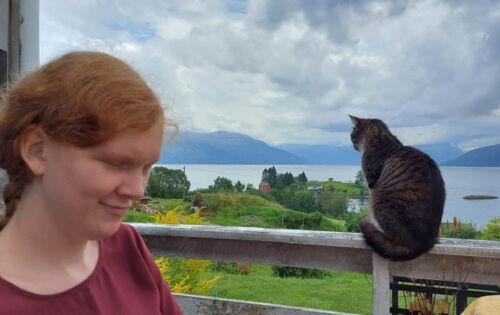 Solveig Marie Oma steht links im Bild und blickt nach unten. Rechts sitzt eine getigerte Katze auf einem Geländer. Im Hintergrund sind ein Fjord, grüne Landschaft, Berge und ein bewölkter Himmel zu sehen.
