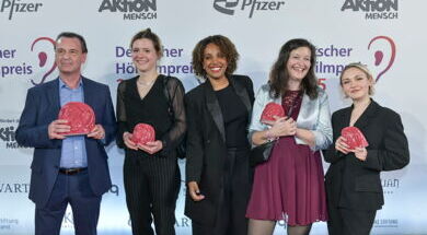 Beim Deutschen Hörfilmpreis steht ein AD-Team steht vor einer Sponsorenwand. Sie halten die Trophäe ADele in ihren Händen. Alle fünf Personen lächeln breit.