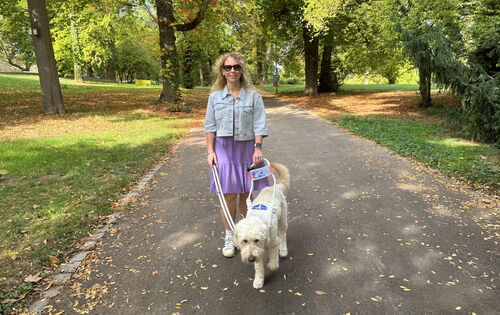 Lisa Mümmler geht mit Führhund Harry im Führgeschirr auf einem Weg im Park, das Ambiente ist herbstlich. Sie trägt Ray-Ban Meta Glasses, eine helle Jeansjacke und ein lilafarbenes Kleid.