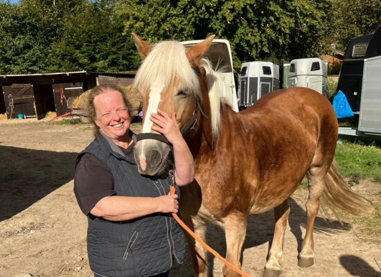 Linda Pröve steht bei einem Haflingerpony, dessen Kopf sie mit einer Hand umfasst. Ihr langes lockiges Haar trägt sie zusammengebunden. Im Hintergrund sind Bäume und Pferdeanhänger zu sehen.