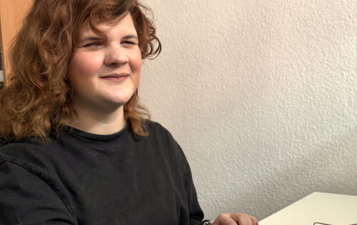 Marie Lampe sitzt an einem Schreibtisch und bedient mit einer Hand die Tastatur, mit der anderen die Braillezeile. Sie hat gewelltes, mittellanges und mitteldunkles Haar.