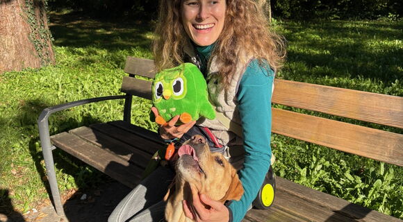 Vivian Hösch sitzt lächelnd im Sonnenschein auf einer Bank. In ihrer rechten Hand hält sie Duo, eine fußballgroße grüne Plüscheule, das Maskottchen von Duolingo. Mit Links streichelt sie ihren hellbraunen Blindenführhund Candy, der neben ihr sitzt.