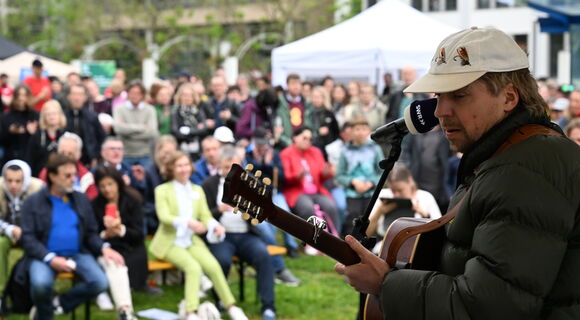 Ein Blick von der Außentribüne auf dem Berliner Platz ins Publikum der Eröffnungsfeier: rechts im Bildvordergrund steht scharf zu sehen Philipp Poisel mit seiner Gitarre, den leicht verschwommenen Besucherinnen und Besuchern zugewandt.