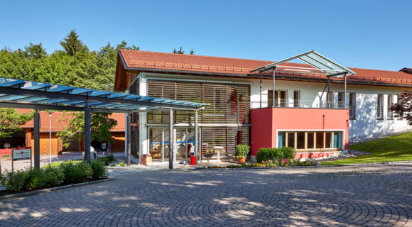 Das Seminargebäude des Aura-Hotels Saulgrub: Die Fassade des Gebäudes ist an zwei Seiten vollverglast. Ein überdachter Weg führt zum Eingang.