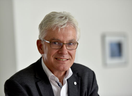 Ein älterer Mann mit weißem Haar, Brille und hellem Teint trägt ein dunkles Sakko über einem weißen Hemd. Er lächelt freundlich und blickt direkt in die Kamera. Der Hintergrund ist neutral und unscharf, mit einem gerahmten Bild an der Wand.