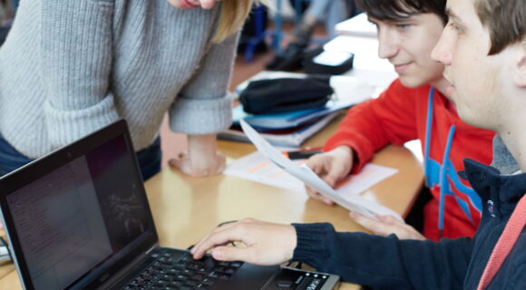 In einem Klassenraum beugt sich eine junge Lehrerin über den Laptop mit Braillezeile eines Schülers. Andere Jugendliche sitzen mit im Raum.