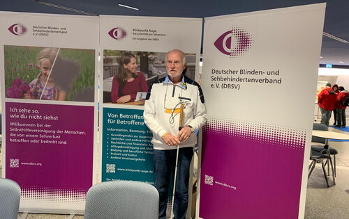Gerd Steting, ein älterer, grauhaariger Herr mit Langstock, steht an einem Messestand. Hinter ihm sind Aufsteller vom DBSV in Purpur und von Blickpunkt Auge in Petrol zu sehen.