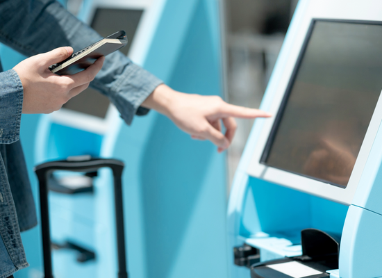 Jemand steht vor einem Automaten und zeigt mit dem Finger darauf. In der anderen Hand hält er ein Handy und ein kleines Heft.