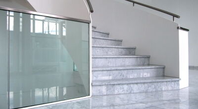 Eine helle Treppe führt in einer Linkskurve nach oben. Sie hat einen silbernen Handlauf und steinerne Stufen.
