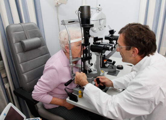 Ein Augenarzt blickt durch ein medizinisches Gerät in die Augen einer älteren Frau, um sie zu untersuchen.