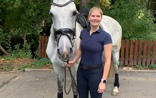 Helen Vogt steht mit ihrer Schimmelstute Janita auf einem Reitplatz, der von Büschen und Bäumen umgeben ist. Sie hat langes blondes, zu einem Pferdeschwanz gebundenes Haar und trägt ein kurzärmeliges Polohemd. Janita trägt einen Sattel und Zaumzeug.