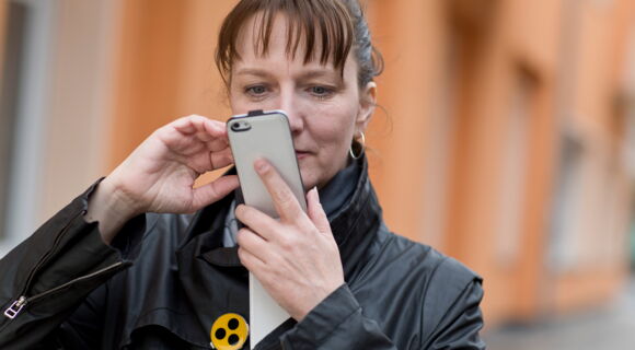 Eine Frau mit Pony-Haarschnitt schaut konzentriert auf ihr Smartphone. An ihrer schwarzen Jacke befindet sich ein gelber Anstecker mit drei Punkten.