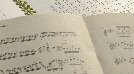 Ein Notenheft in Schwarzschrift liegt auf einem Notenheft in Braille. Jemand hat mit einem Bleistift etwas darauf notiert.