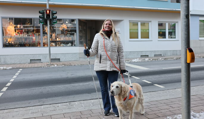In der rechten Hand ihren Langstock, in der linken den Führbügel ihres hellen Führhunds: So steht Mandy Kugel in Steppjacke und mit Handschuhen auf der Straße vor einem Haus.