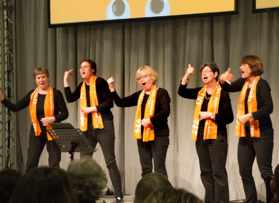 Gebärdenchor: Fünf Frauen in schwarzer Kleidung und mit dem orangefarbenen Kirchentagsschal vom Kirchentag 2017 in Berlin und Wittenberg auf einer Bühne. Mit der rechten Hand machen sie eine Geste mit ausgestrecktem Daumen und kleinem Finger.