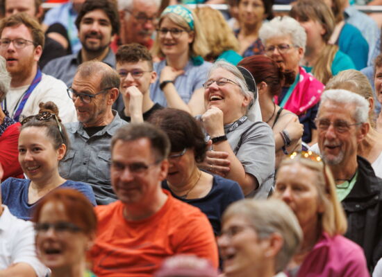 Bild eines Publikums beim Kirchentag 2023 in Nürnberg: Lachende und lächelnde Menschen verschiedenen Alters blicken in eine Richtung. Einige tragen kurzärmelige T-Shirts.