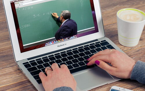 Eine Person tippt auf einem MacBook, auf dessen Bildschirm ein Video mit einem Lehrer läuft, der an eine grüne Tafel schreibt. Die Person trägt graue Kleidung, hat lila lackierte Nägel und trägt Ringe. Daneben stehen eine Tasse Kaffee und ein Smartpho