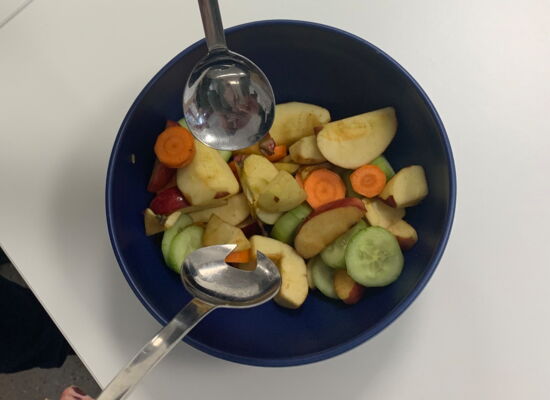 Ein bunter Salat wird in einer dunklen Schüssel mit Salatbesteck vermengt. Das Gefäß hebt sich kontrastreich von der hellen Arbeitsplatte ab.