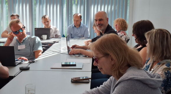 Beim Workshop „Überregionale Kooperation“: Teilnehmerinnen und Teilnehmer sitzen an Tischen. Vor manchen liegt ein Smartphone, andere haben ihr Laptop aufgeklappt. Auch Workshop-Leiter Reiner Delgado bedient ein Laptop, während er lächelnd Richtung Kamera