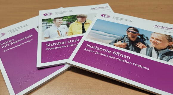 Drei Ausgaben der DBSV-Jahrespublikation "Weitersehen" liegen aufgefächert auf einem Tisch. "Leben mit Sehverlust", "Sichtbar stark" und "Horizonte öffnen" steht weiß auf purpur auf dem jeweiligen Heft.