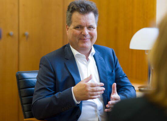 Porträt von Jürgen Dusel. Er trägt ein blaues Jacket und hält die Hände beim Sprechen ausgebreitet.