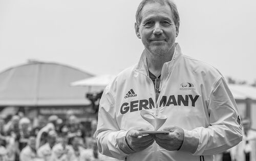 Dr. Karl Quade mit einer Trophäe bei den Paralympics in Rio de Janeiro. Er trägt kurzes, schütteres Haar und hat einen Dreitagebart. Er lächelt in die Kamera. Auf seiner hellen Jacke steht „GERMANY“.