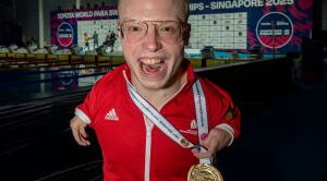 Josia Topf freut sich über seine Goldmedaille. Er ist ein bonder Mann mit TAR-Syndrom, und hat sehr kurze Arme. Er hat den Mund freudig geöffnet und strahlt in die Kamera.