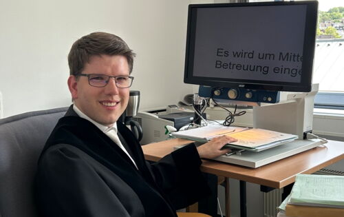 André Stahl sitzt in Richterrobe am Schreibtisch seines Büros. Er hat kurzes dunkleres Haar und trägt eine Brille. Sein Monitor zeigt einen stark vergrößerten Text an.