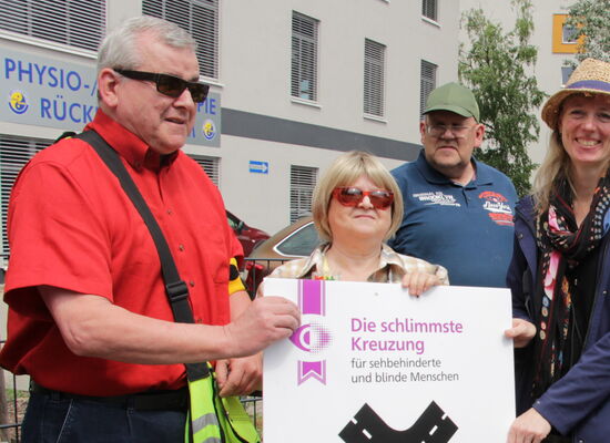 Auf einem Gehweg: Bodo Rinas (links) und Margrit Richter (2.v.l.) übergeben die plakatgroße Urkunde zur schlimmsten Kreuzung an die Behindertenbeauftragte von Königs Wusterhausen, Friederike Weigelt (mittig). Weitere Beteiligte schauen zu.