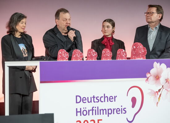 Klaus-Jörg Kaminski, blinder AD-Autor des Films „Paris, Texas“ spricht in ein Mikrofon bei der Deutschen Hörfilm-Verleihung. Neben ihm stehen sein Team und die Laudatorin des Films. Auf einem Tisch stehen sechs rosafarbene Hörfilmpreis-Trophäen.