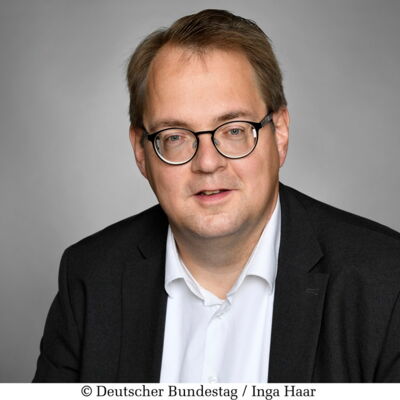 Sören Pellmann mit Brille und hellem Teint trägt einen dunklen Anzug mit weißem Hemd. Er blickt freundlich in die Kamera vor neutralem Hintergrund.