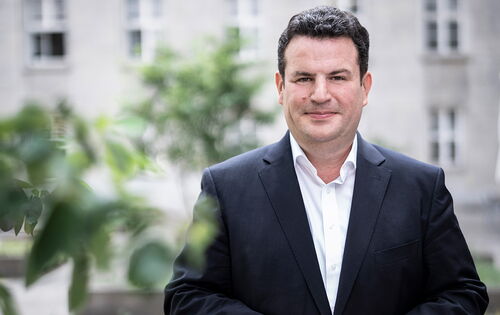 Bundesarbeitsminister Hubertus Heil schaut lächelnd direkt in die Kamera. Er trägt einen dunklen Sakko mit einem weißen Hemd. Er hat schwarze Haare, sein Bart ist kurz rasiert. Im Hintergrund sind helle Gebäude und Pflanzen zu sehen.