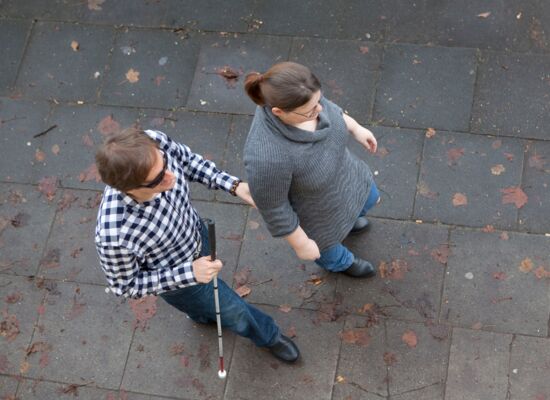 Zwei Menschen gehen eine Straße entlang. Von oben ist zu erkennen, dass sie etwas versetzt hintereinandergehen. Dabei berührt der hintere Mann den Ellenbogen der vorne gehenden Frau. Beide tragen Jeans und grau/schwarze Oberteile.