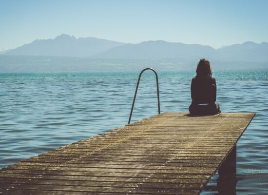 Am Ende eines Holzstegs sitzt eine Frau und blickt mit abgewandtem Rücken aufs Meer.