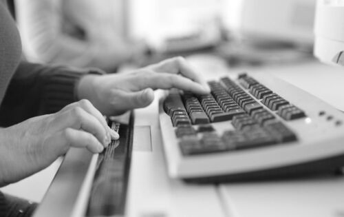 Frauenhände an einer Computertastatur mit Braillezeile. Im Hintergrund ist ein weiterer Arbeitsplatz zu sehen.