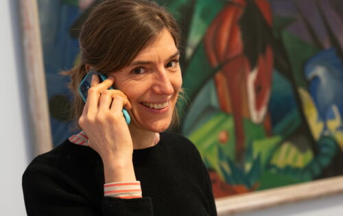 Eine Frau mit glatten, braunen Haaren hält sich lächelnd ein Smartphone ans Ohr. Hinter ihr ist ein abstraktes Gemälde zu erkennen.