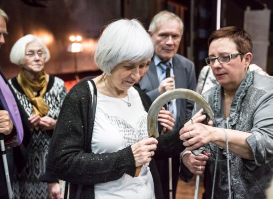 Bei einer Theaterführung betastet eine ältere Frau eine große Sichel. Sie lächelt. Eine Frau rechts von ihr scheint sie dabei zu unterstützen. Rund herum stehen fünf weitere Menschen, einige von ihnen mit Langstock.