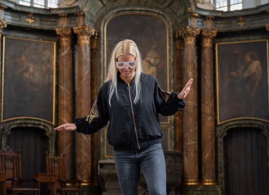 Eine junge Frau mit Simulationsbrille geht in einer Kirche drei nicht markierte Stufen hinunter. Unsicher balanciert sie mit den Armen.