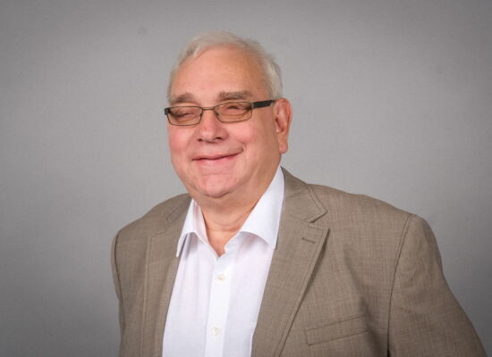 Portrait Hans-Werner Lange. Er trägt ein weißes Hemd, ein hellbeiges Sakko und eine dunkle, getönte Brille. Er hat kurzes weißes Haar.