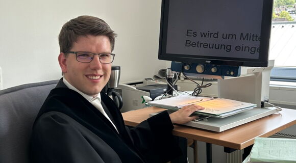 André Stahl sitzt in Richterrobe am Schreibtisch seines Büros. Er hat kurzes dunkleres Haar und trägt eine Brille. Sein Monitor zeigt einen stark vergrößerten Text an.