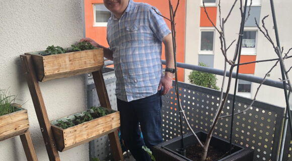 Rainer Heim trägt ein helles, kariertes Hemd und steht lächelnd auf seinem Balkon. Neben ihm ein dreistufiges Hochbeet mit Kräutern, vor ihm einige Blumenkübel mit wachsenden Pflanzen.