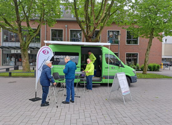 Das Beratungsmobil des Blinden- und Sehbehindertenvereins Westfalen ist   auf einem Platz geparkt. Johannes Willenberg steht vor dem Wagen an einem  Stehtisch und blickt zu zwei älteren Herren, die sich dem Fahrzeug nähern.