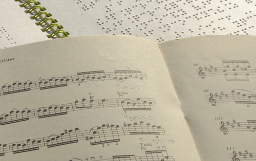 Ein Notenheft in Schwarzschrift liegt auf einem Notenheft in Braille. Jemand hat mit einem Bleistift etwas darauf notiert.