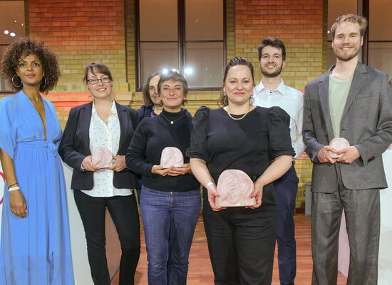 In festlicher Kleidung steht das AD-Team des Animationsfilms „Arielle, die Meerjungfrau“ auf der Bühne. Es gewann die ADele in der Kategorie „Spielfilm - TV/Mediatheken/Streaming“.  Einige Team-Mtglieder halten die Keramik-Trophäe in den Händen.