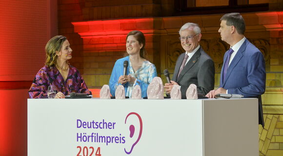 Auf der Bühne hinter einem weißen Pult mit dem Logo des Deutschen Hörfilmpreises stehen folgende Personen von links nach rechts: Nadine Heidenreich, Dr. Ulrike Köhler, Bundespräsident a. D. Prof. Dr. Horst Köhler und Dr. Thomas Krämer