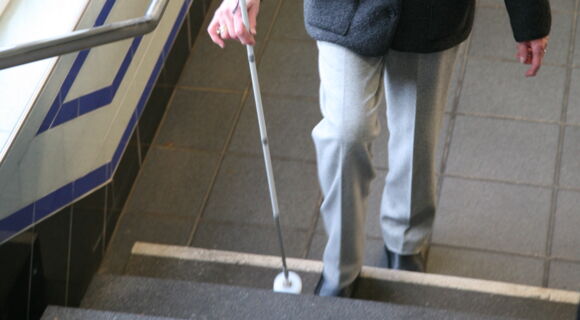 Eine ältere Frau mit Langstock steigt eine Treppe hinauf. Die unterste Stufe ist deutlich visuell gekennzeichnet.
