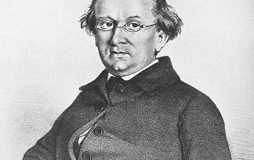 Zeichnung eines Mannes, Eduard Mörike, mit Brille und gewelltem Haar. Er trägt einen dunklen Mantel mit mehreren Knöpfen und sitzt mit leicht geneigtem Kopf. Der Hintergrund ist schlicht.