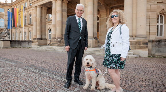 Vor dem Neuen Schloss in Stuttgart stehen von links nach rechts Ministerpräsident Winfried Kretschmann, Festivalführhund Harry und Sichtweisen-Redakteurin Lisa Mümmler.